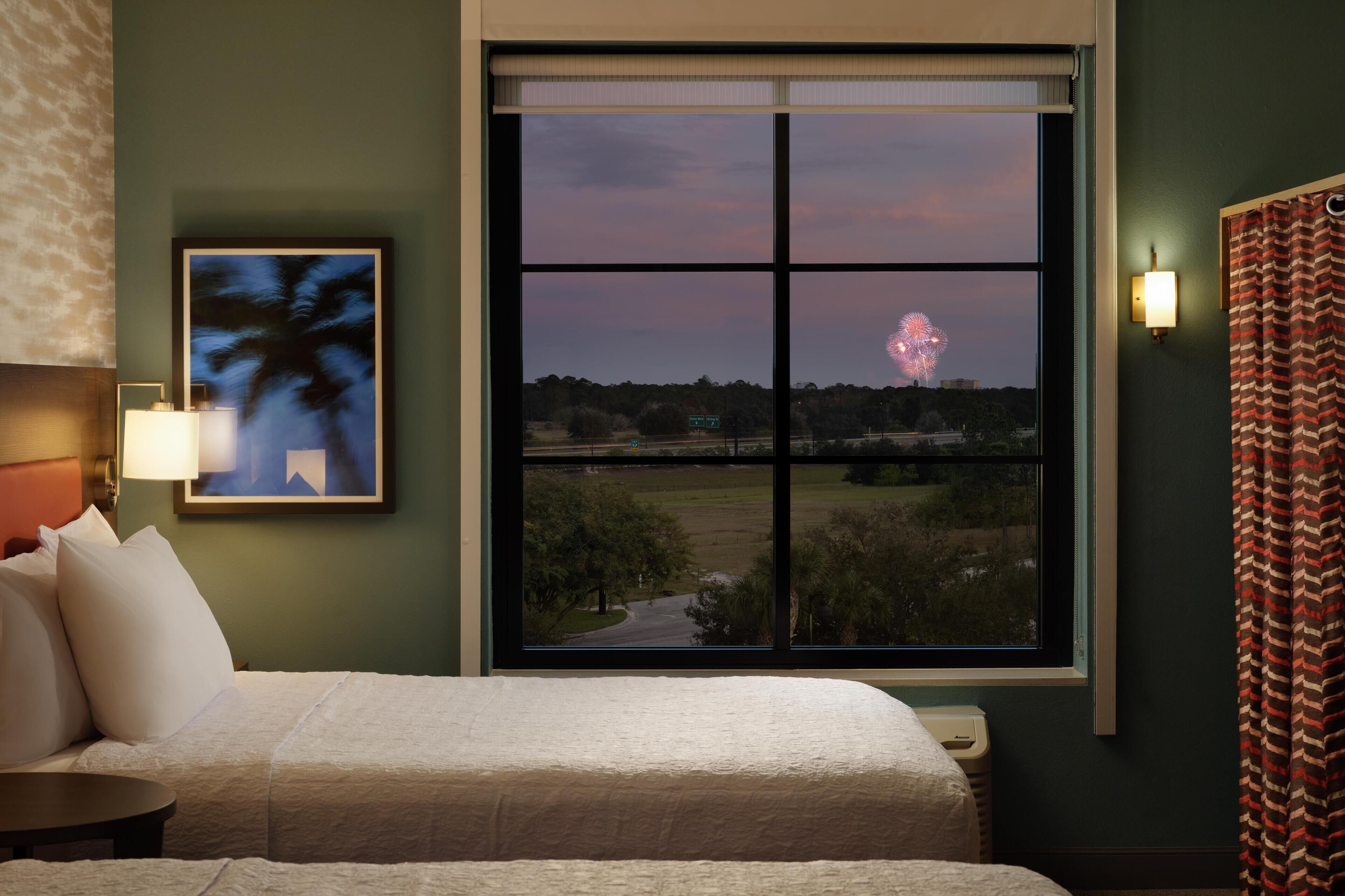 A hotel room with a large window, a bed, a nightstand, a lamp, and a framed picture on the wall.