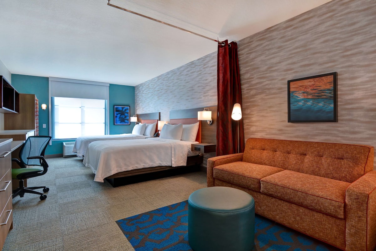 Habitación de hotel con una cama, dos mesitas de noche, un escritorio con una silla, un sofá, un otoman redondo y un cuadro en la pared.