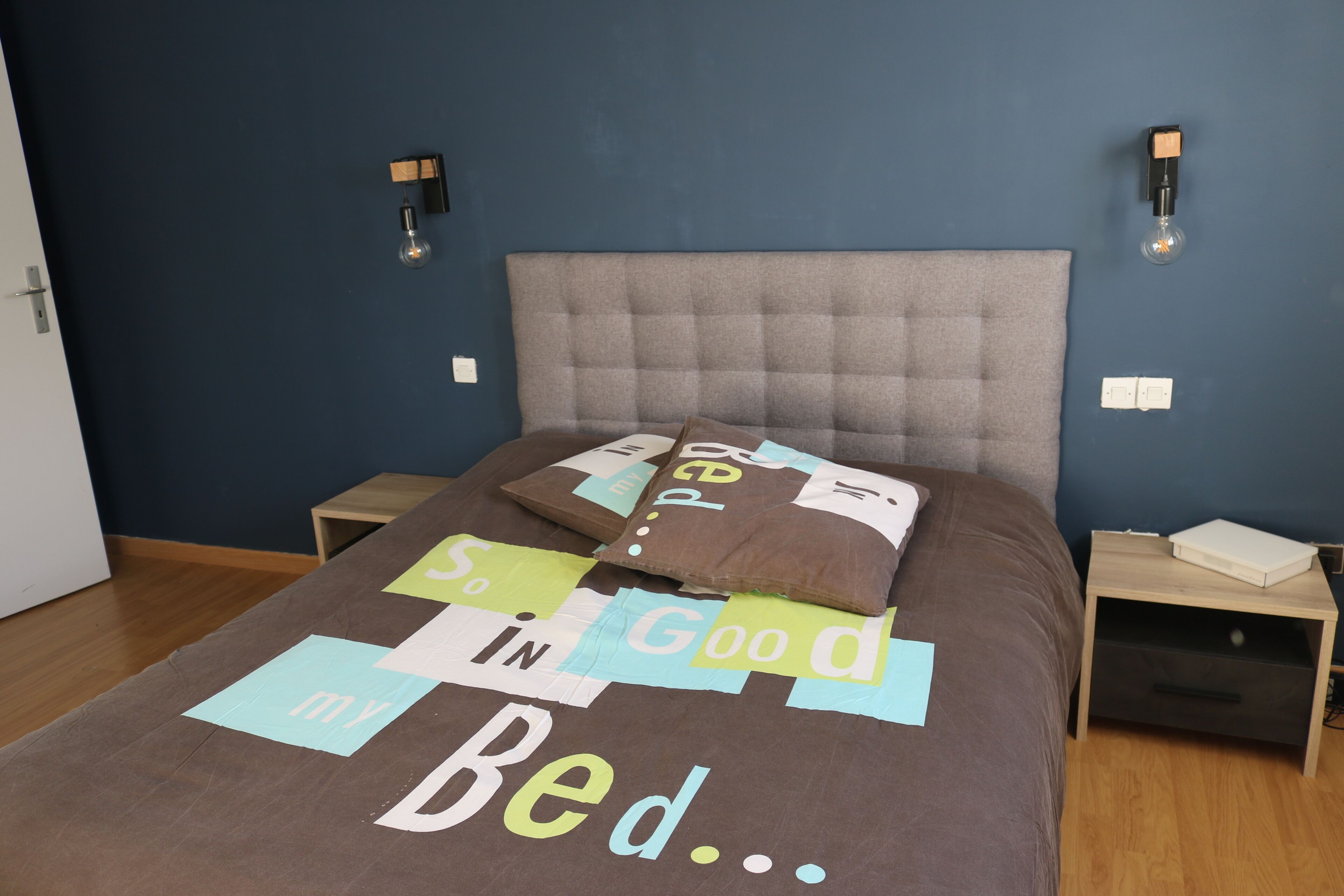 Een net opgemaakt bed met een kleurrijk sprei, twee nachtkastjes en wandlampen.