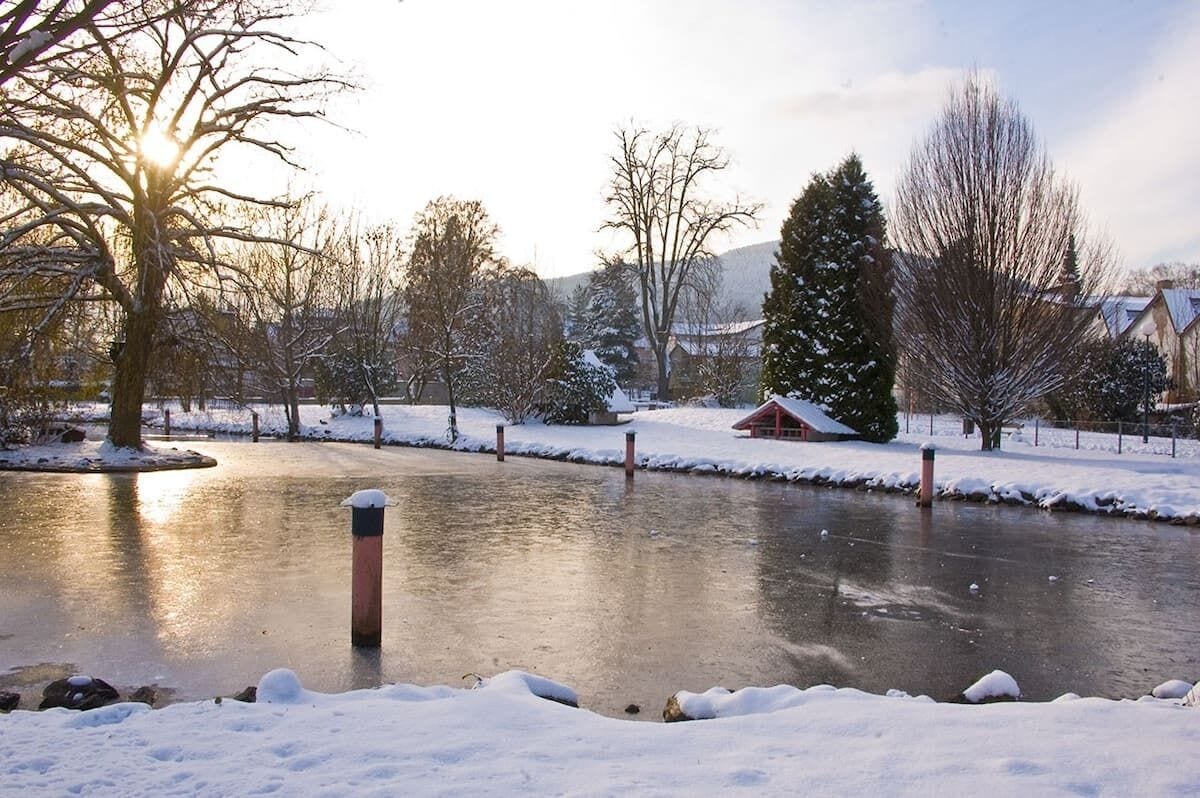 Une rivière gelée aux rives et aux arbres couverts de neige, un bâtiment rouge et un ciel dégagé.