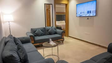 Une chambre d’hôtel moderne, avec un canapé gris, une petite table basse, un téléviseur à écran plat fixé au mur et une porte en bois donnant sur une chambre à coucher.