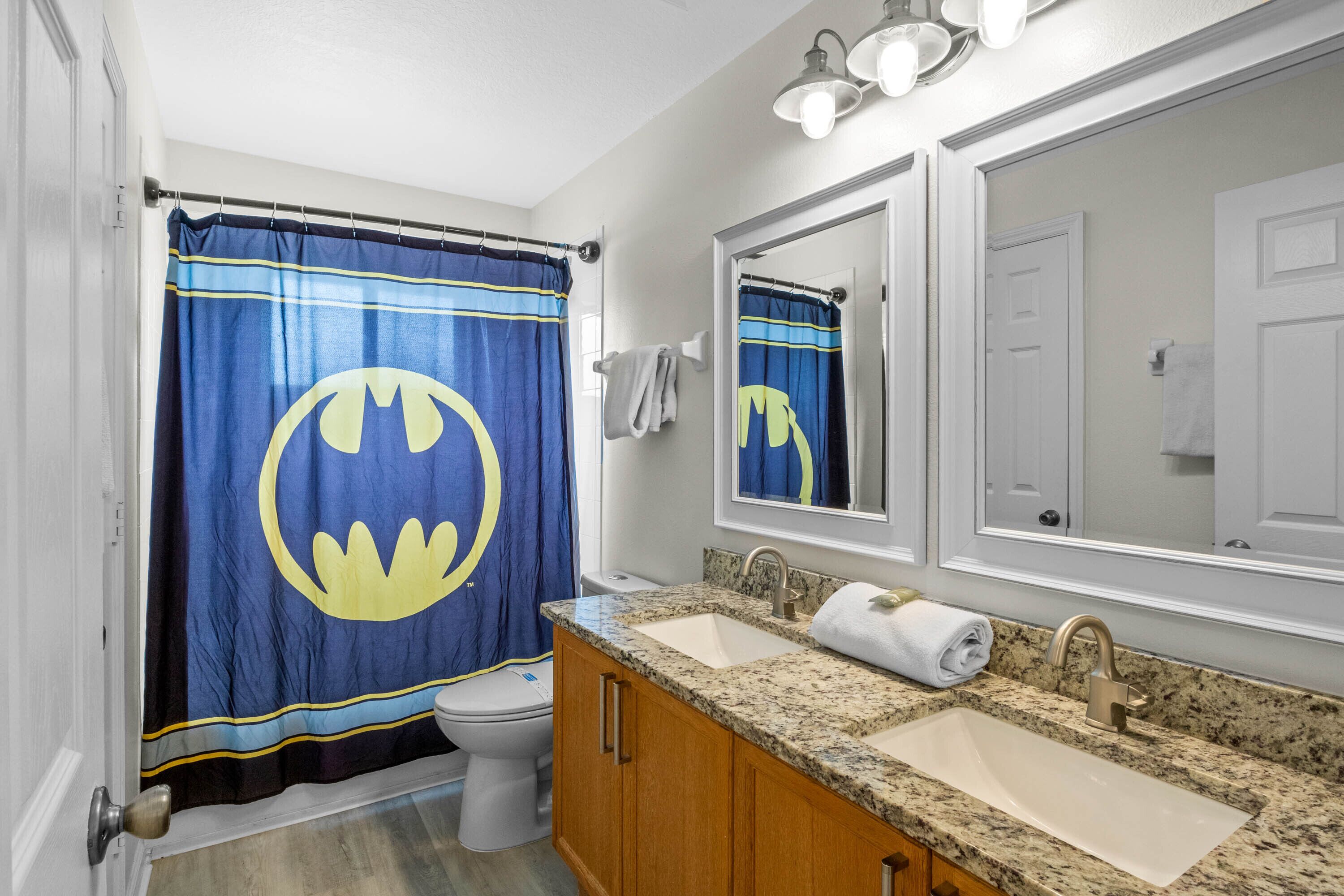 Een badkamer met een Batman-thema douchegordijn, een wit toilet, een wastafel met een opbergmeubel en een grote spiegel erboven.