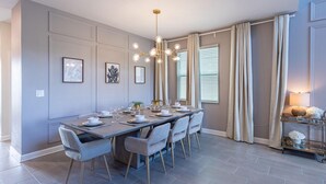 Un coin repas avec une longue table dressée pour six personnes, des murs de couleur gris clair, un lustre et une fenêtre avec des rideaux.