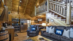 Interior de una cabaña de troncos con cocina, comedor y una sala de estar acogedora con mobiliario de cuero y chimenea.