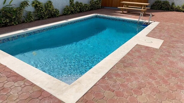 A rectangular swimming pool with a tiled edge, surrounded by a brick patio and a white wall with greenery. - Beautiful Bahamas Unit 4 (Nassau)