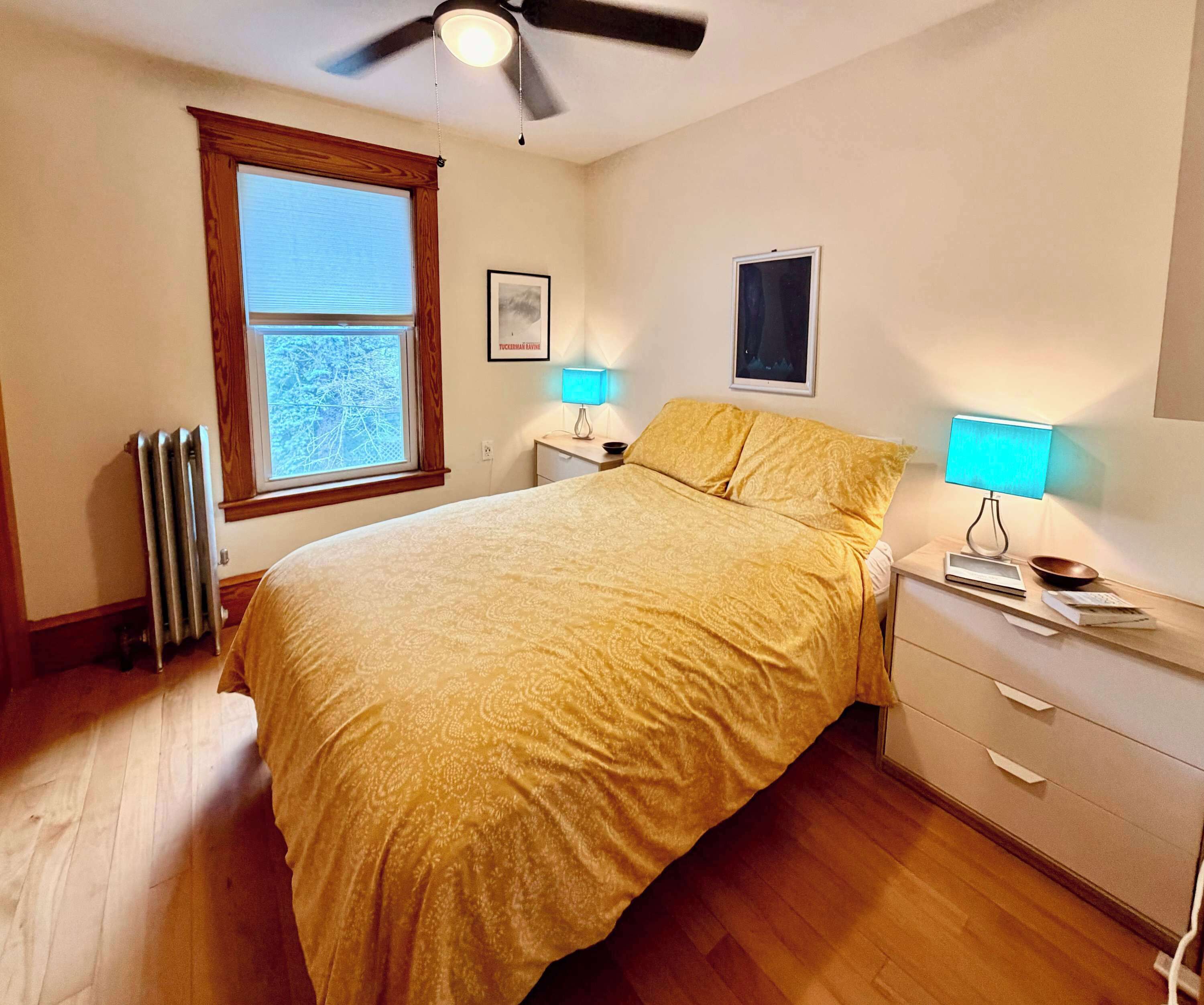 A bedroom with a bed, two bedside tables, a ceiling fan, a window with blinds, and a radiator.