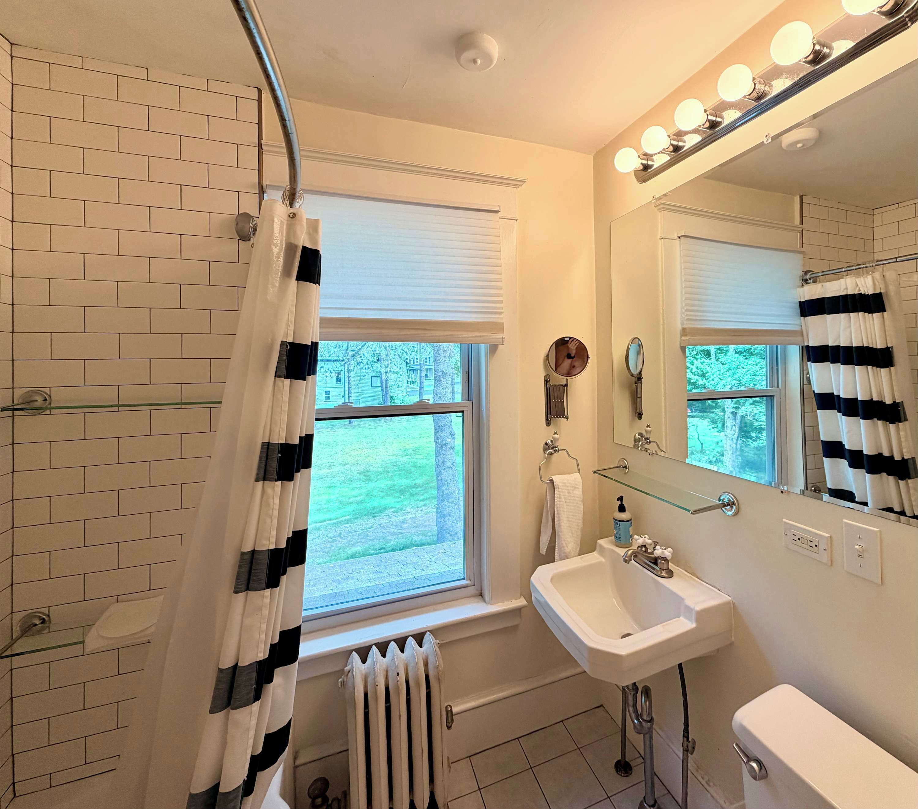 Ein Badezimmer mit Dusche, Waschbecken, Toilette, einem beleuchteten Spiegel und einem Fenster mit Jalousien.
