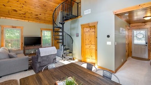 A cozy living room with a wooden ceiling, a spiral staircase, a grey sofa, a wooden dining table, and a TV.