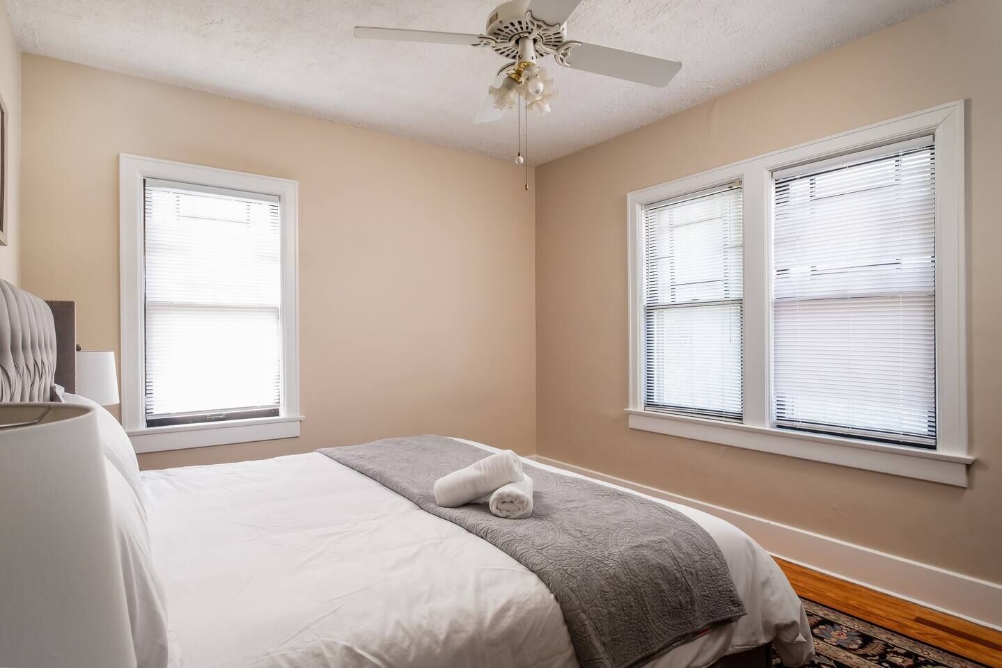 Une chambre à coucher avec un lit, deux fenêtres équipées de stores, un ventilateur de plafond et un tapis.