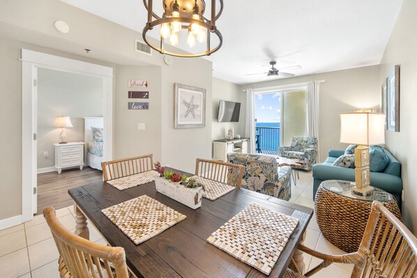A spacious living room with a large wooden dining table, comfortable seating, a balcony with a view, and a ceiling fan.