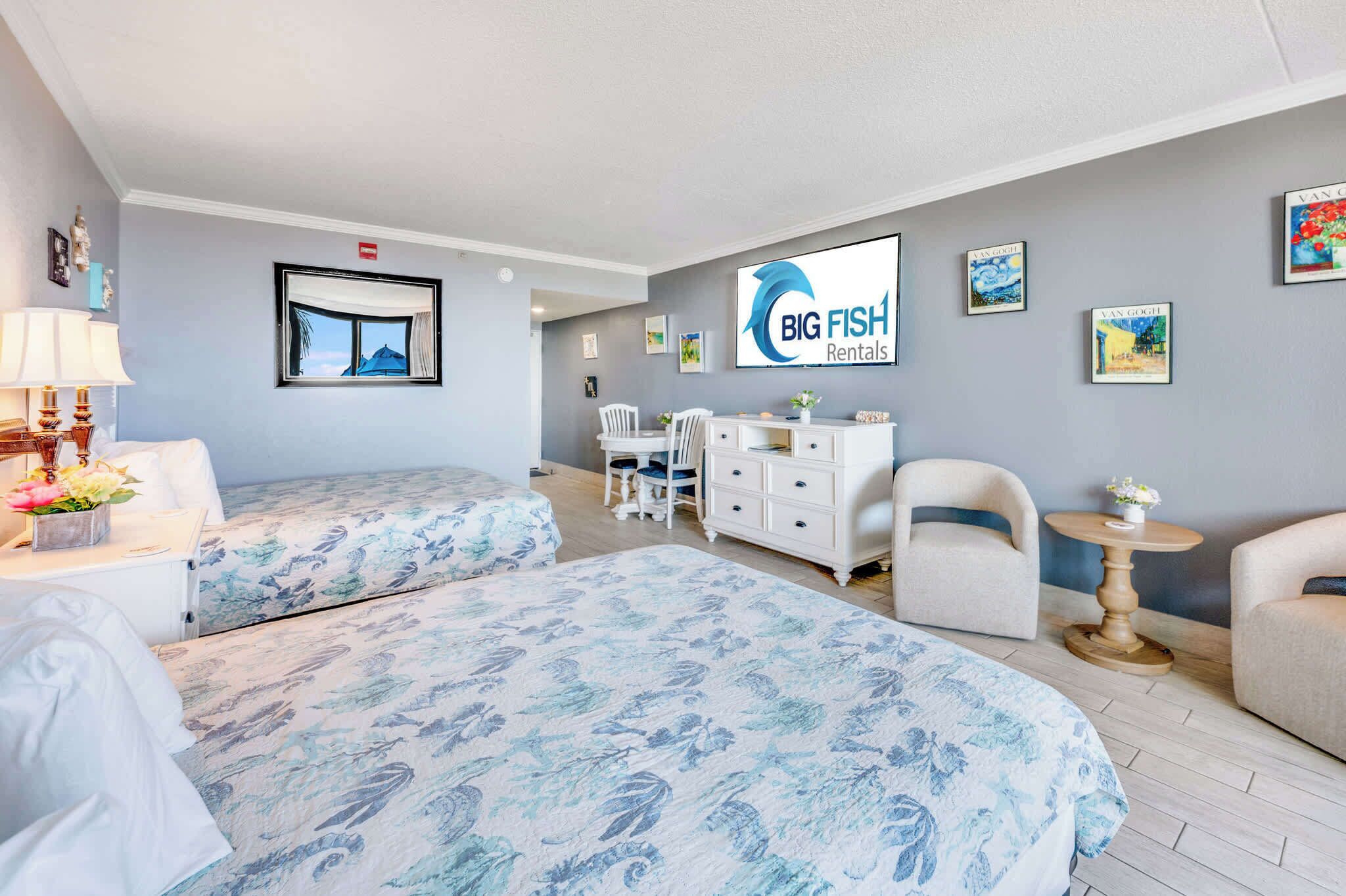 A hotel room with two beds, a desk, a chair, a small table, and artwork on the walls.