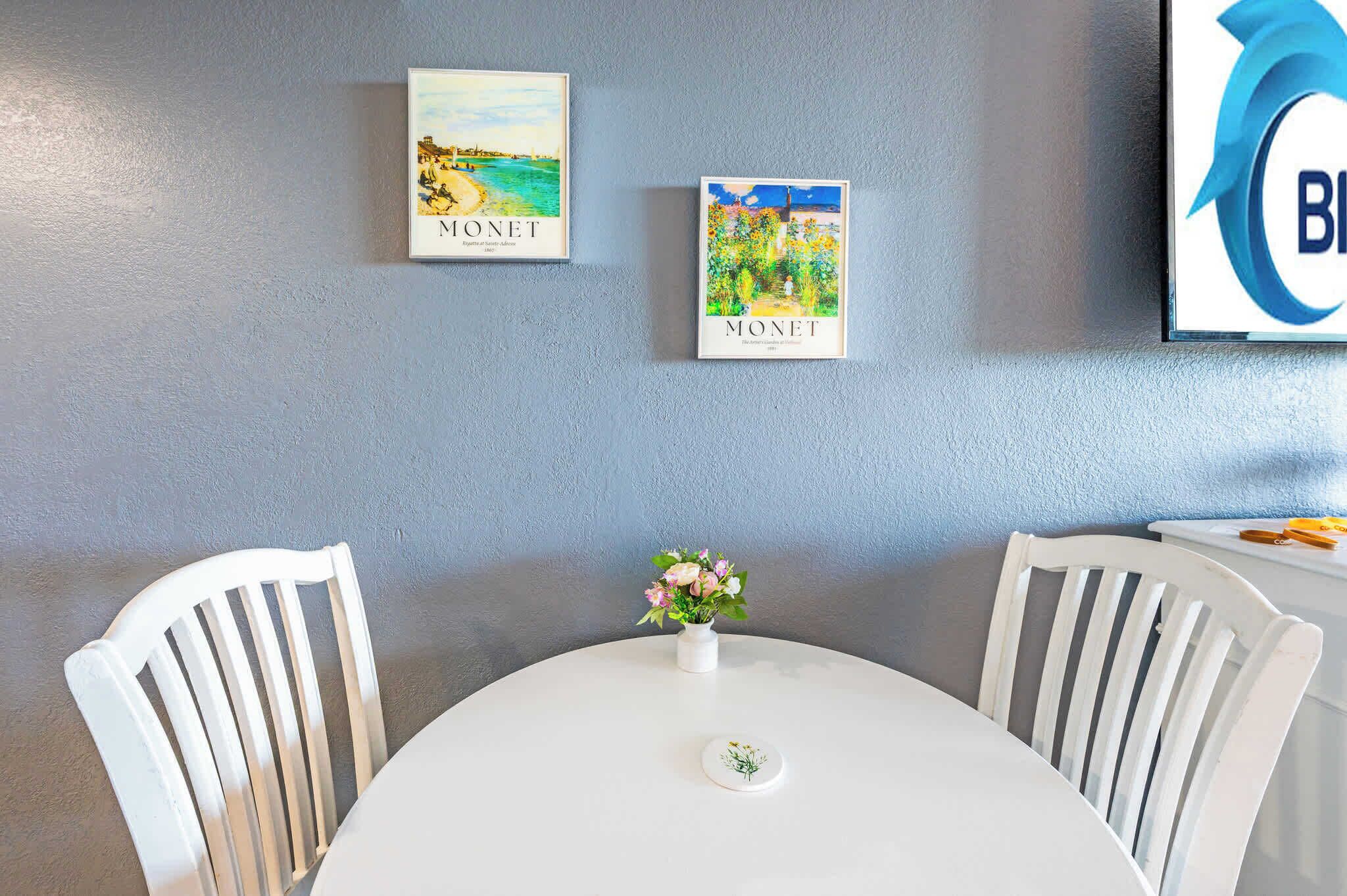 A dining area with a round table, white chairs, a vase with flowers, and two Monet paintings on the wall.