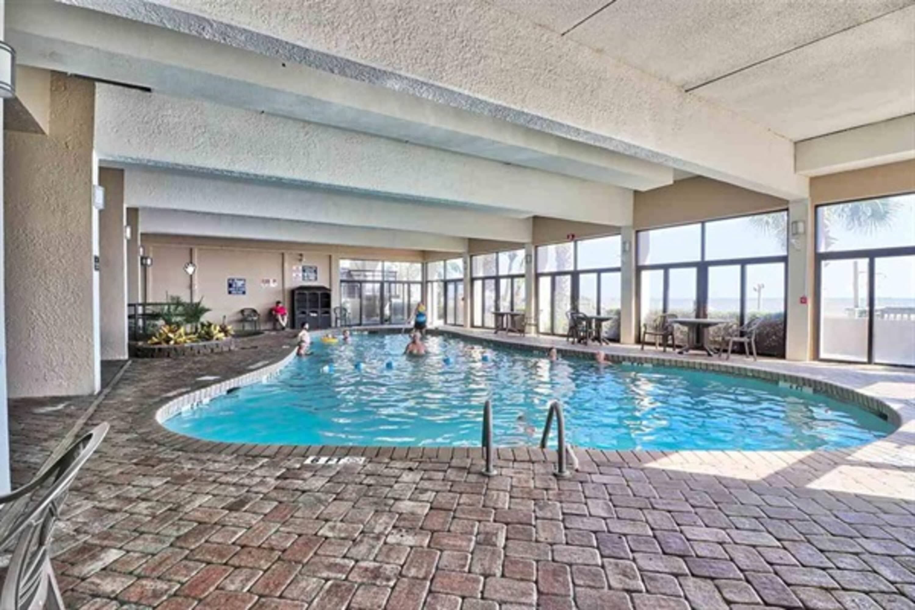 An indoor swimming pool with clear glass walls, a brick floor, and a view of the outdoors.