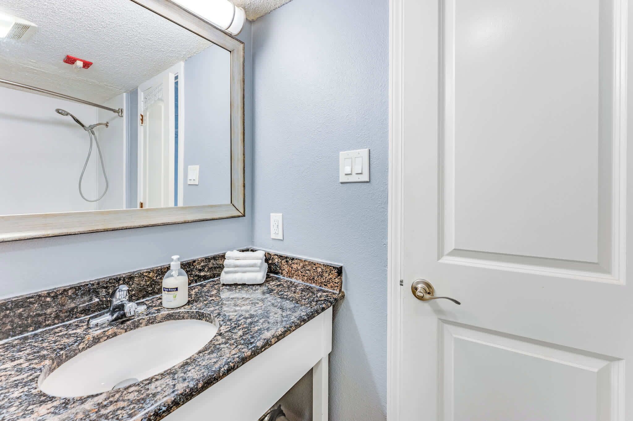 A bathroom with a granite countertop sink, a mirror, a shower, and a white door.