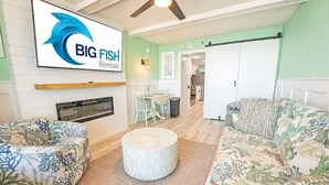 A spacious living room with two patterned sofas, a round coffee table, and a dining area with a white table and chairs. A large TV screen displays a logo. - Amazing  Beachfront Views, Decorated and Renovated (Myrtle Beach)