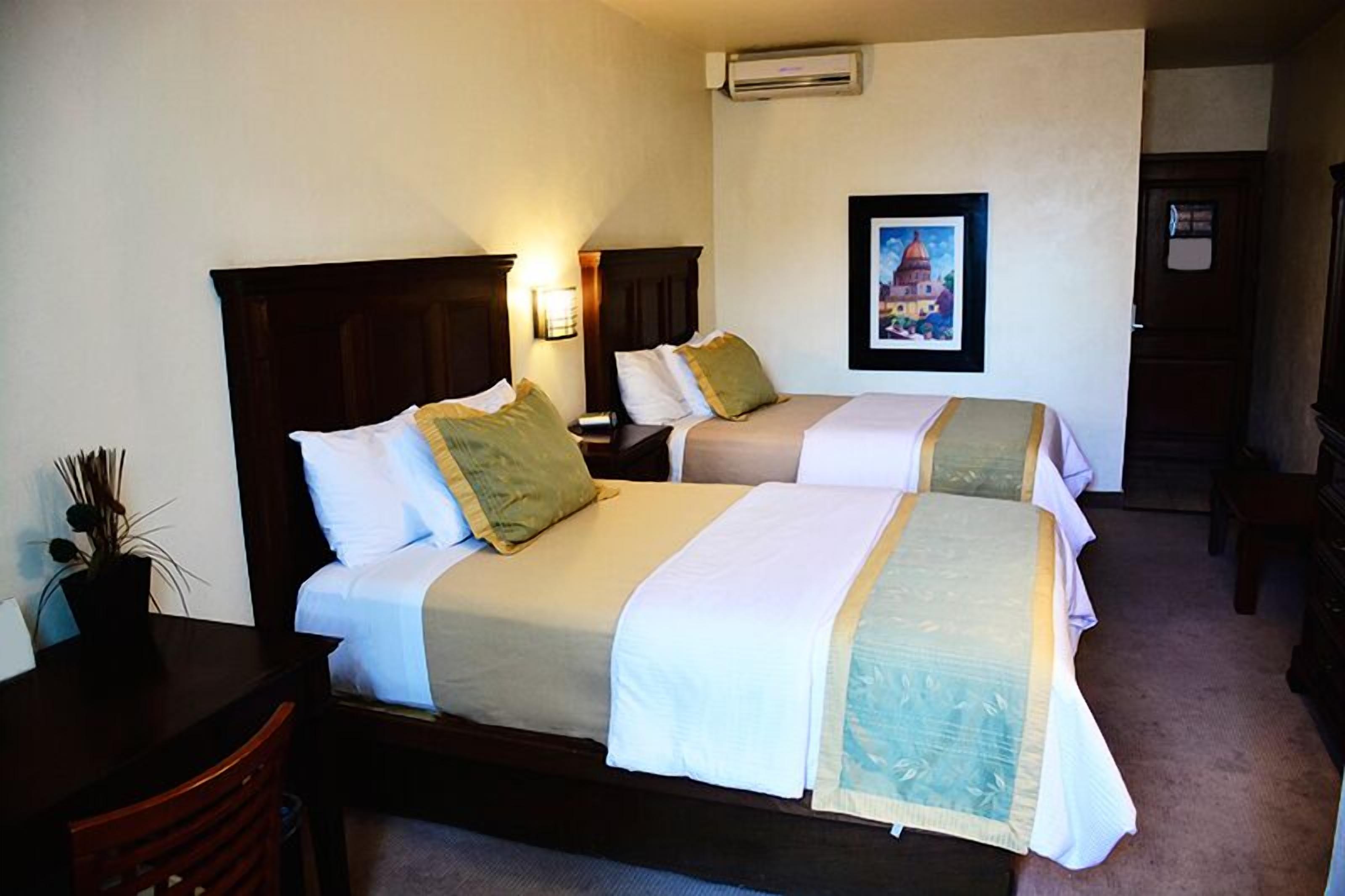 Ein Hotelzimmer mit zwei Einzelbetten, einem Schreibtisch mit Stuhl, einem Blumentopf und einem gerahmten Bild an der Wand.