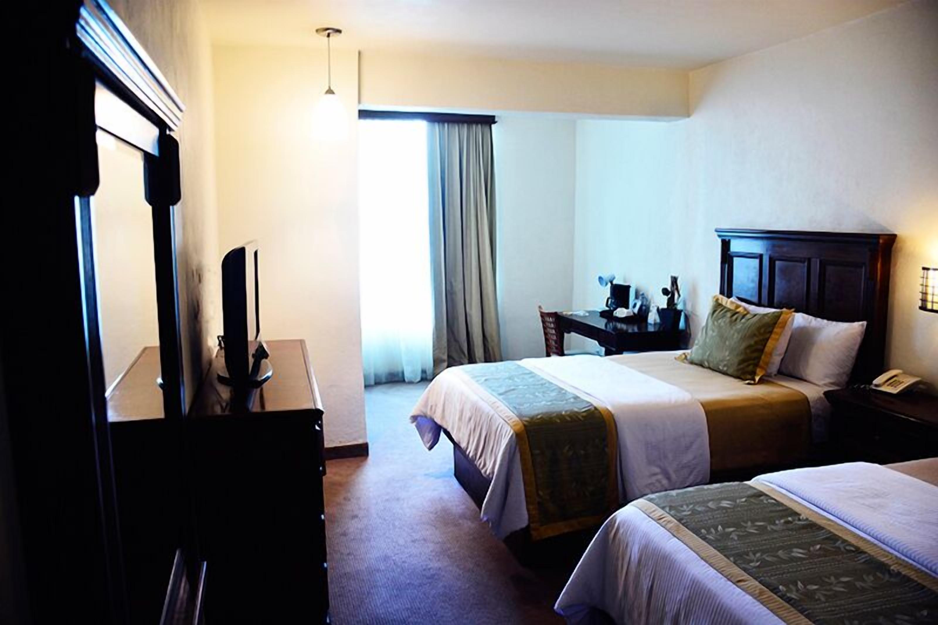 Habitación de hotel con dos camas, un escritorio con silla, un aparador y un ventanal con cortinas.