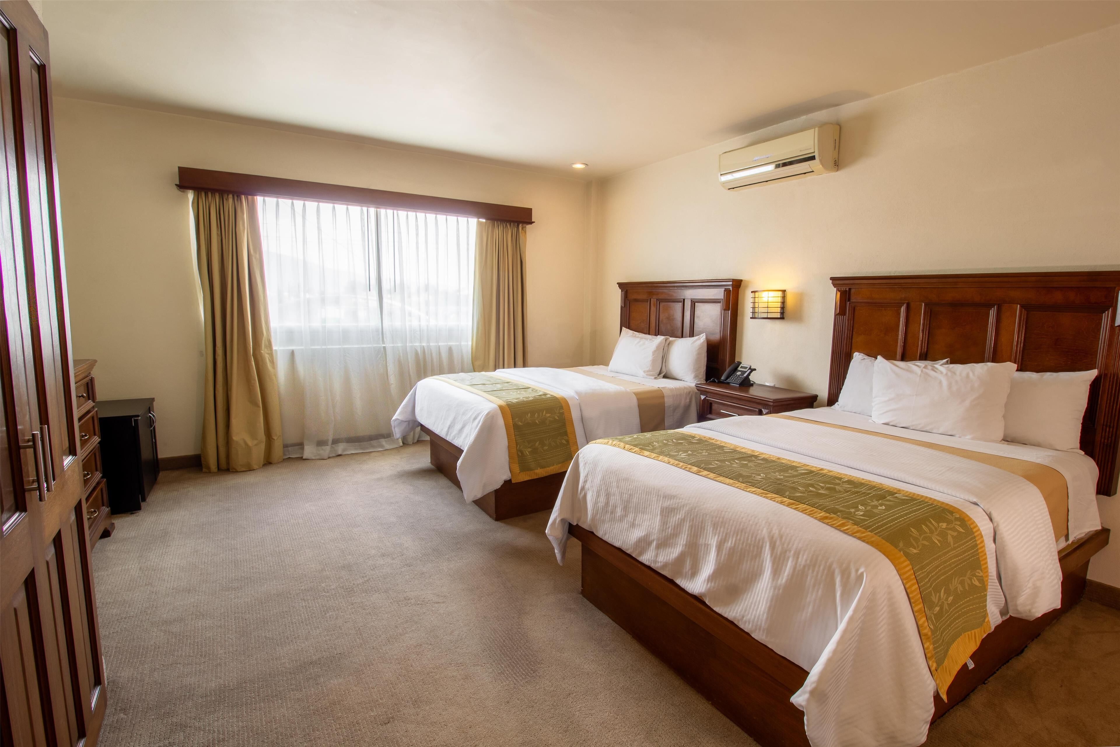 A hotel room with two beds, a wooden wardrobe, an air conditioning unit, and a window with curtains.