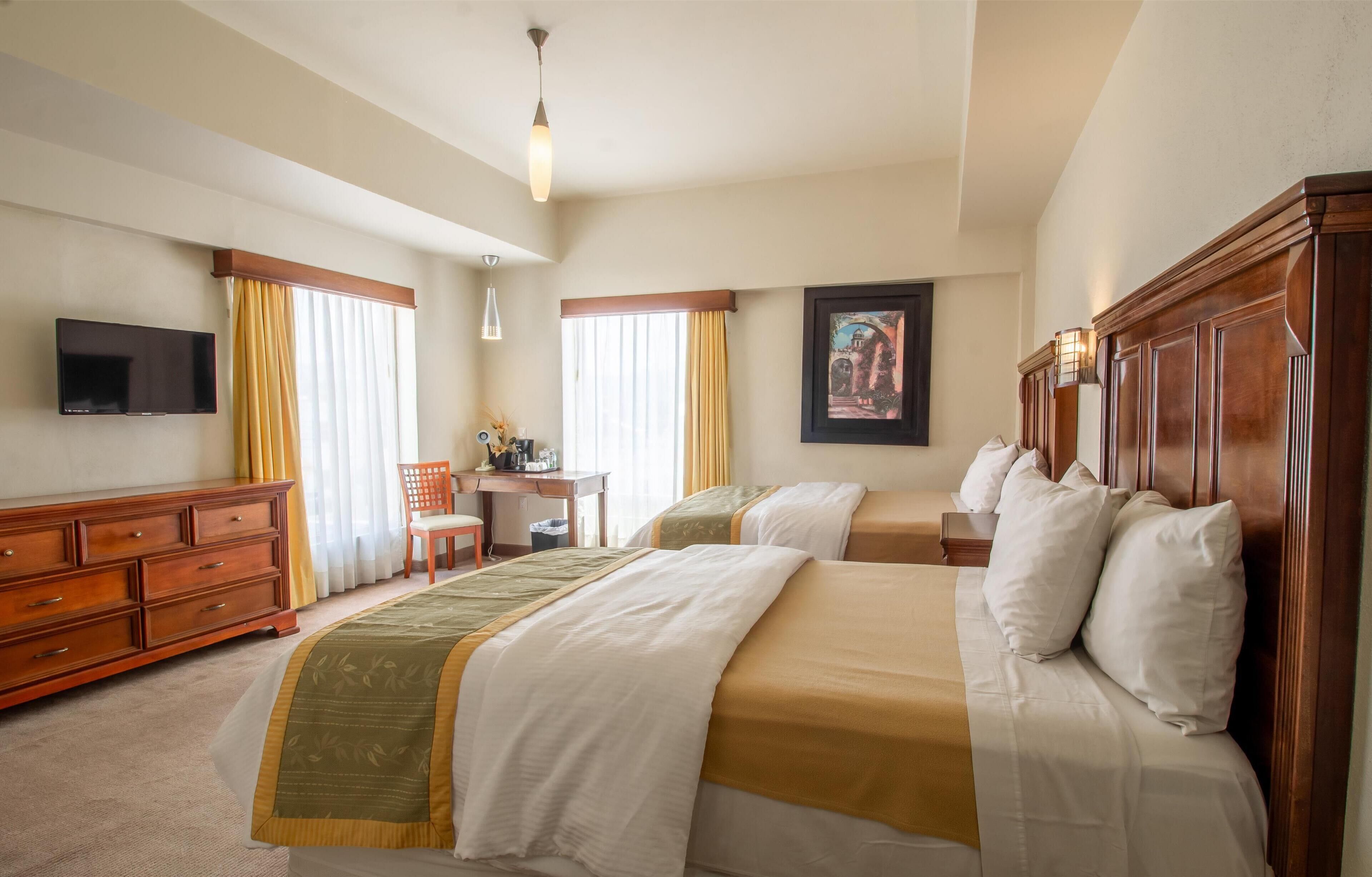 A hotel room with two beds, a desk, a chair, a TV, and a painting on the wall.