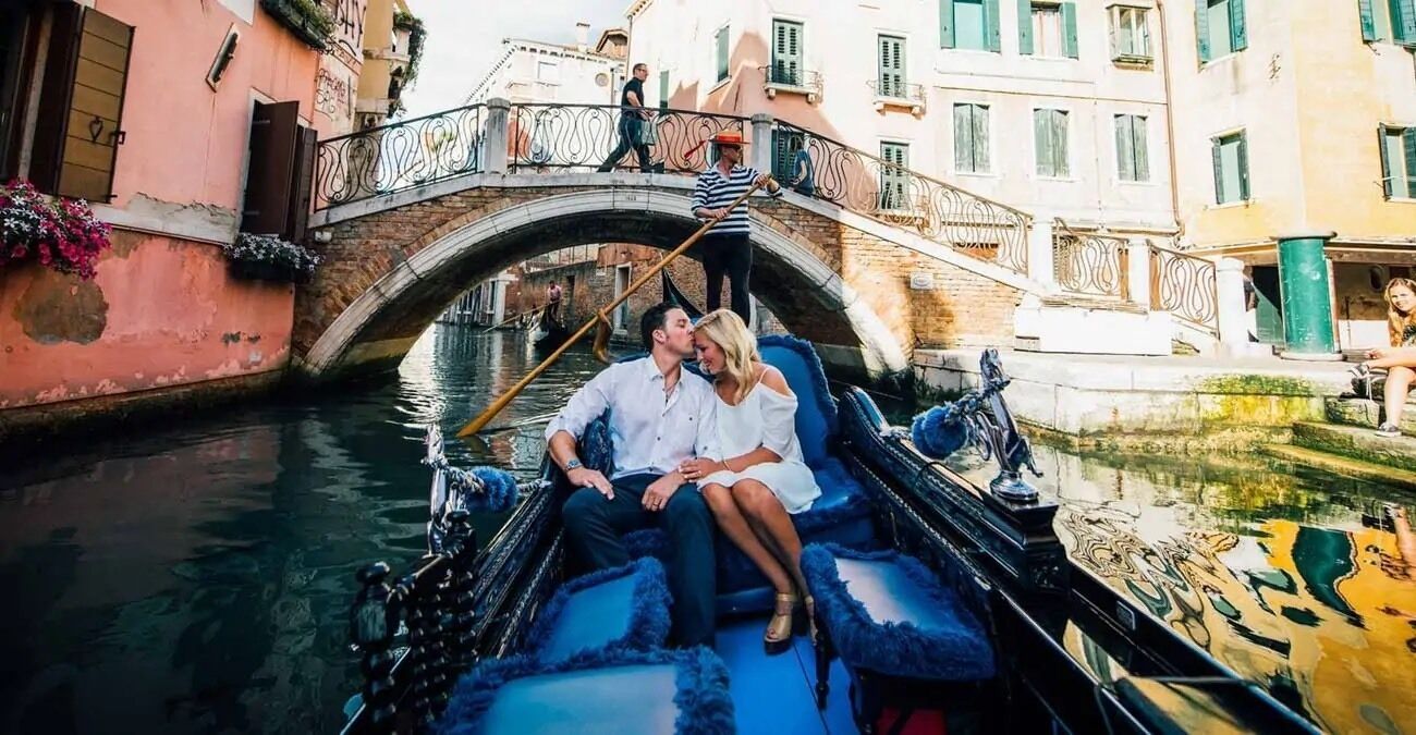 Un bateau de plaisance sur un canal, un couple assis à l’intérieur, un pont en arrière-plan et des bâtiments le long du canal.