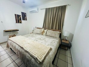 A bedroom with a bed, a bedside table with a lamp, a wall-mounted air conditioner, and a window with curtains. - Charming Condo - Walk to the Beach, Dinning & Nightlife (Cabo San Lucas)