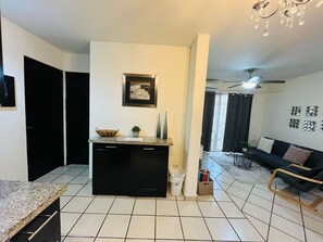 A modern kitchen with black cabinets, a granite countertop, and a small island. There is a framed picture on the wall, a trash can, and a basket of books. The living area has a dark sofa, a small table, and a chandelier.