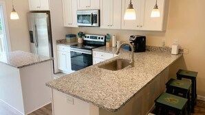 A kitchen with white cabinets, a stainless steel refrigerator, a microwave, a stove, and a sink. There are three stools and pendant lights above the counter. - Beach Haven (saint augustine)