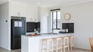 A modern kitchen with white cabinets, a black refrigerator, a window with blinds, a wooden clock, and a bar area with stools.