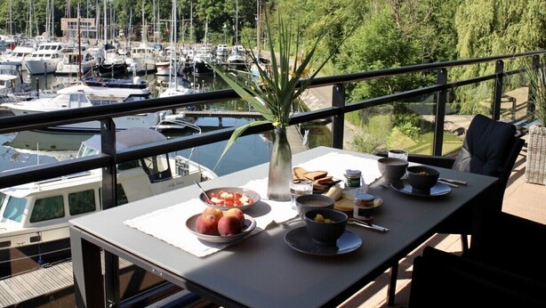 Une table à manger avec des aliments pour le petit-déjeuner et un vase de fleurs, donnant sur une marina où sont amarrés des bateaux.
