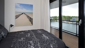 Une chambre à coucher comprenant un lit, une table de chevet, une lampe, un tableau représentant une plage, et un balcon avec vue sur une étendue d’eau et de la verdure.