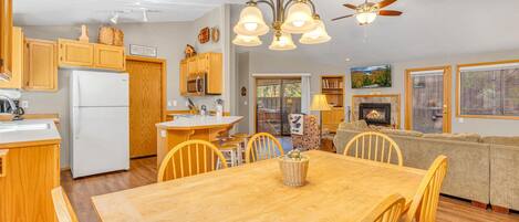A cozy cabin interior with a wooden dining table, chairs, a kitchen with wooden cabinets, a fireplace, and a ceiling fan.