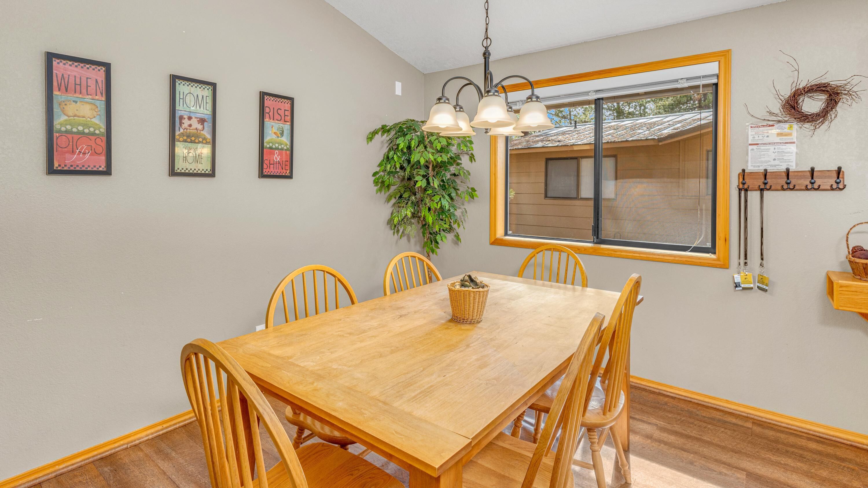A dining room with a wooden table and chairs, framed artwork on the wall, a hanging plant, and a window with a view of the outside.