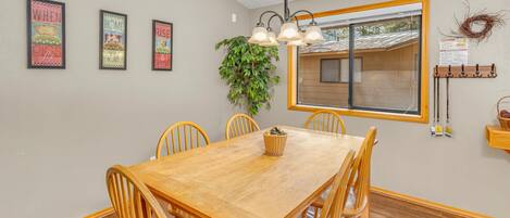 A dining room with a wooden table and chairs, framed artwork on the wall, a hanging plant, and a window with a view of the outside.