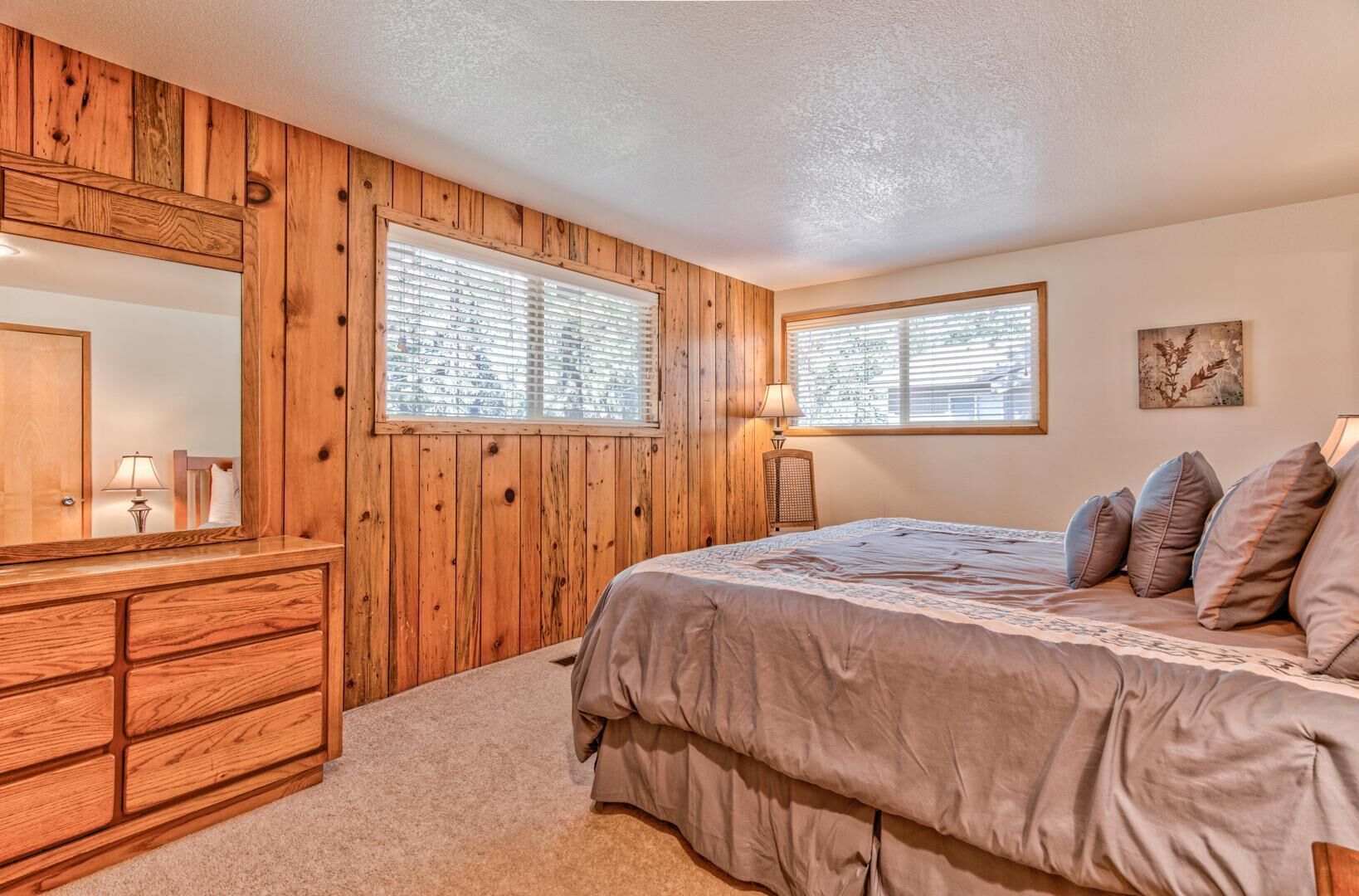 A bedroom with wooden panel walls, a bed with pillows, a dresser, and two windows with blinds.