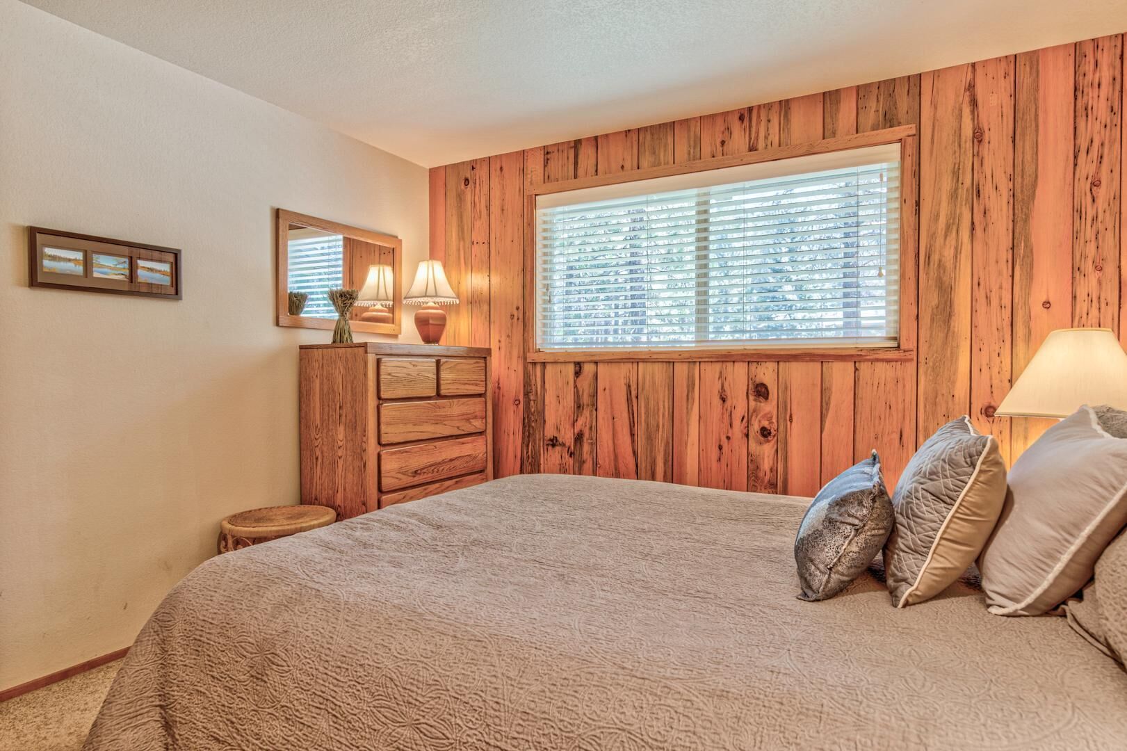 A bedroom with a wooden paneled wall, a large window with blinds, a bed with a patterned bedspread, two bedside lamps, and a small wooden stool.