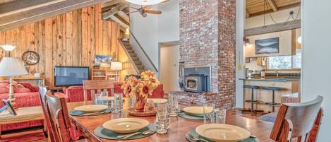 A dining area with a wooden table set for four, a brick fireplace, and a kitchen in the background.