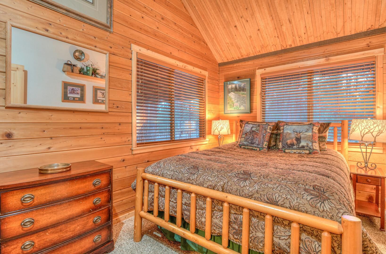 A cozy log cabin bedroom with a wooden bed, two bedside tables, a dresser, and a window with blinds.