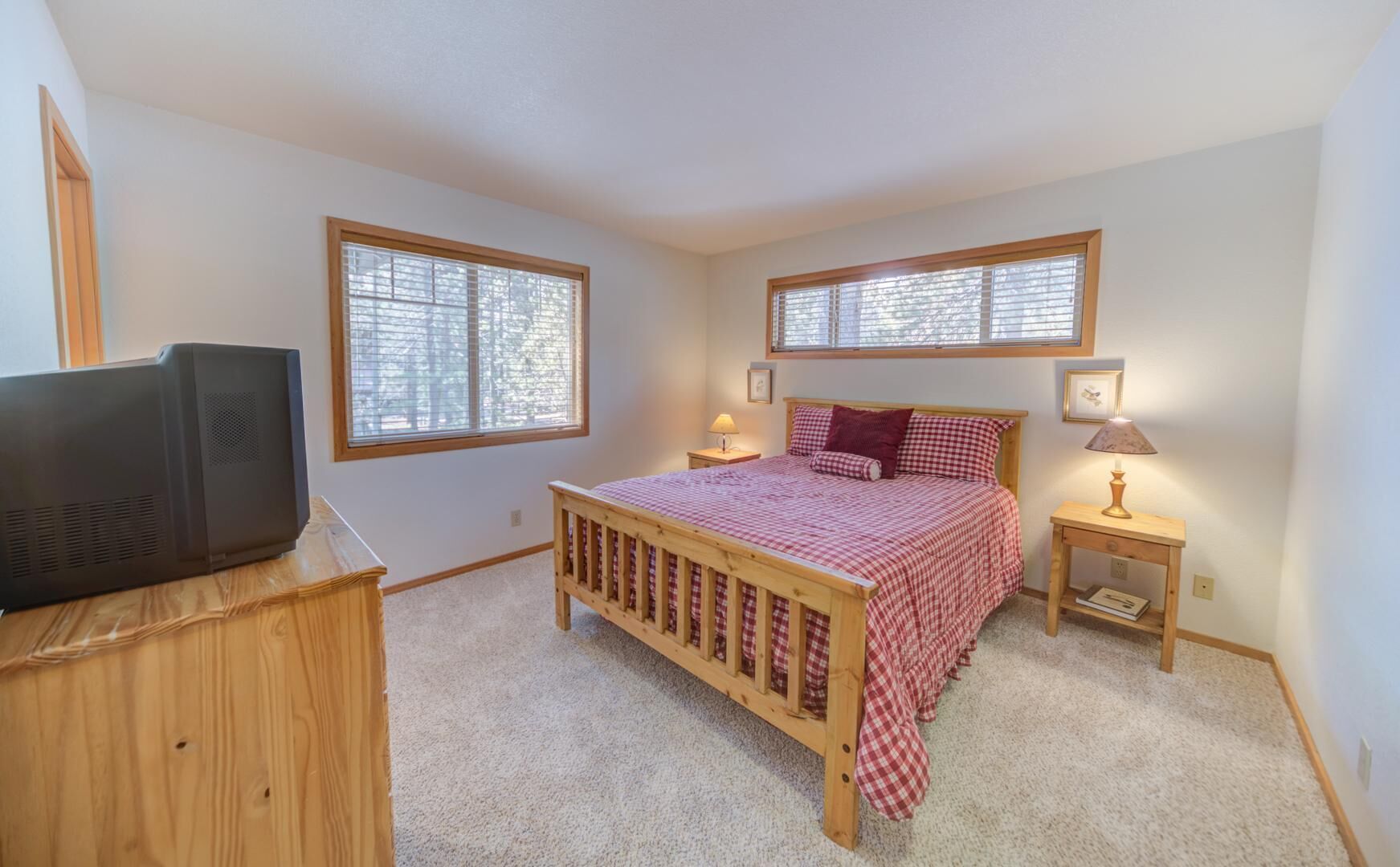 A bedroom with a wooden bed, two bedside tables with lamps, a television on a wooden cabinet, and large windows with blinds.