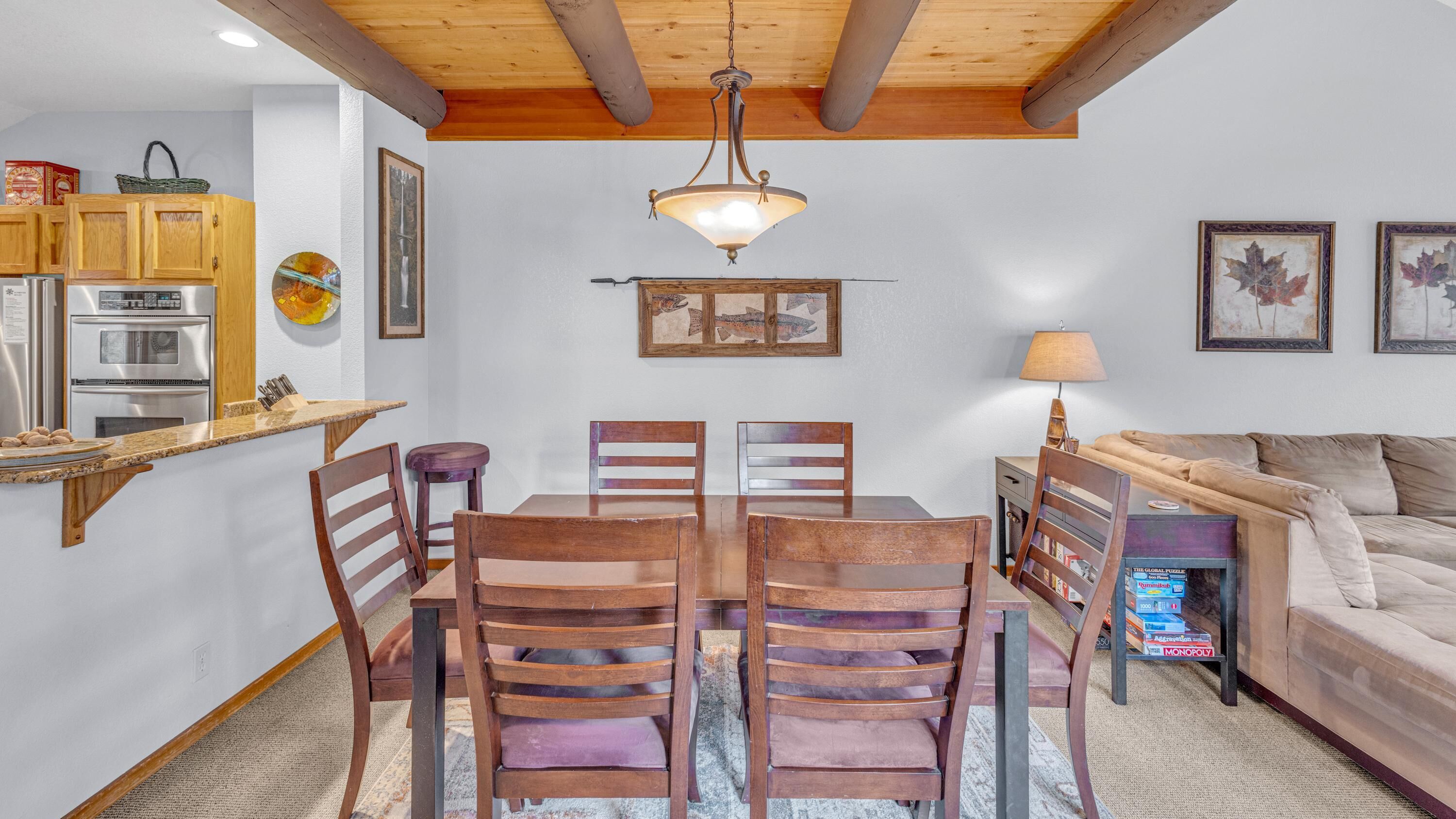 Un comedor con una mesa y sillas de madera, una araña y una cocina al fondo.