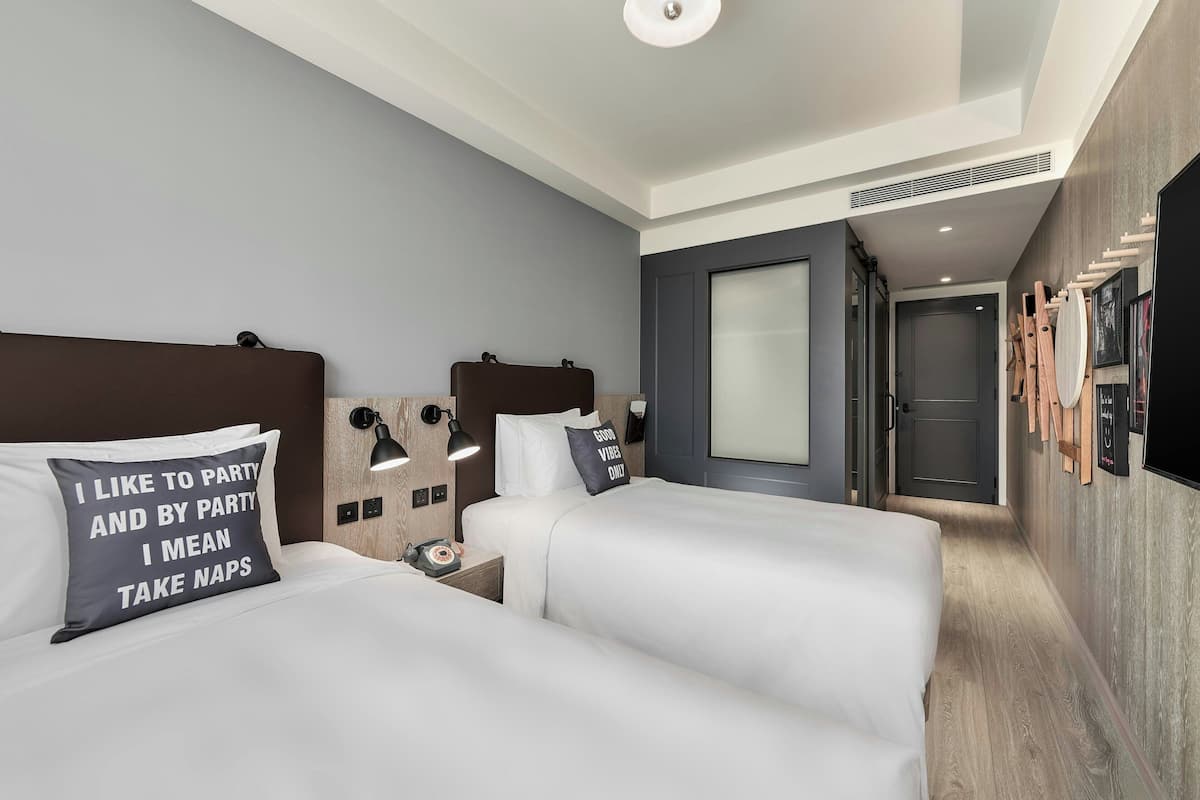 A hotel room with two beds, a desk, a lamp, and a TV.