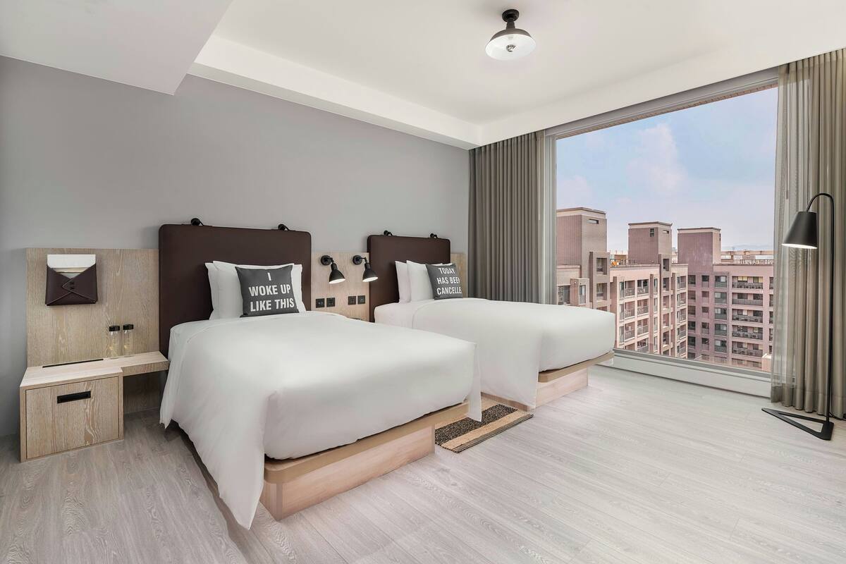 A modern hotel room with two beds, a large window with a city view, a ceiling light, and a bedside table with a lamp.