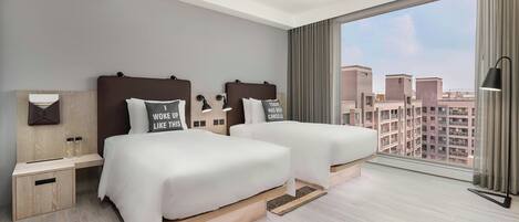 A modern hotel room with two beds, a large window with a city view, a ceiling light, and a bedside table with a lamp.