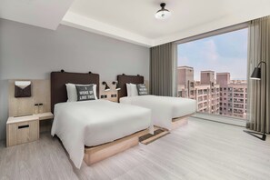 Quarto de hotel moderno com duas camas, uma janela ampla com vista para a cidade, uma luminária no teto e uma mesa de cabeceira com abajur.
