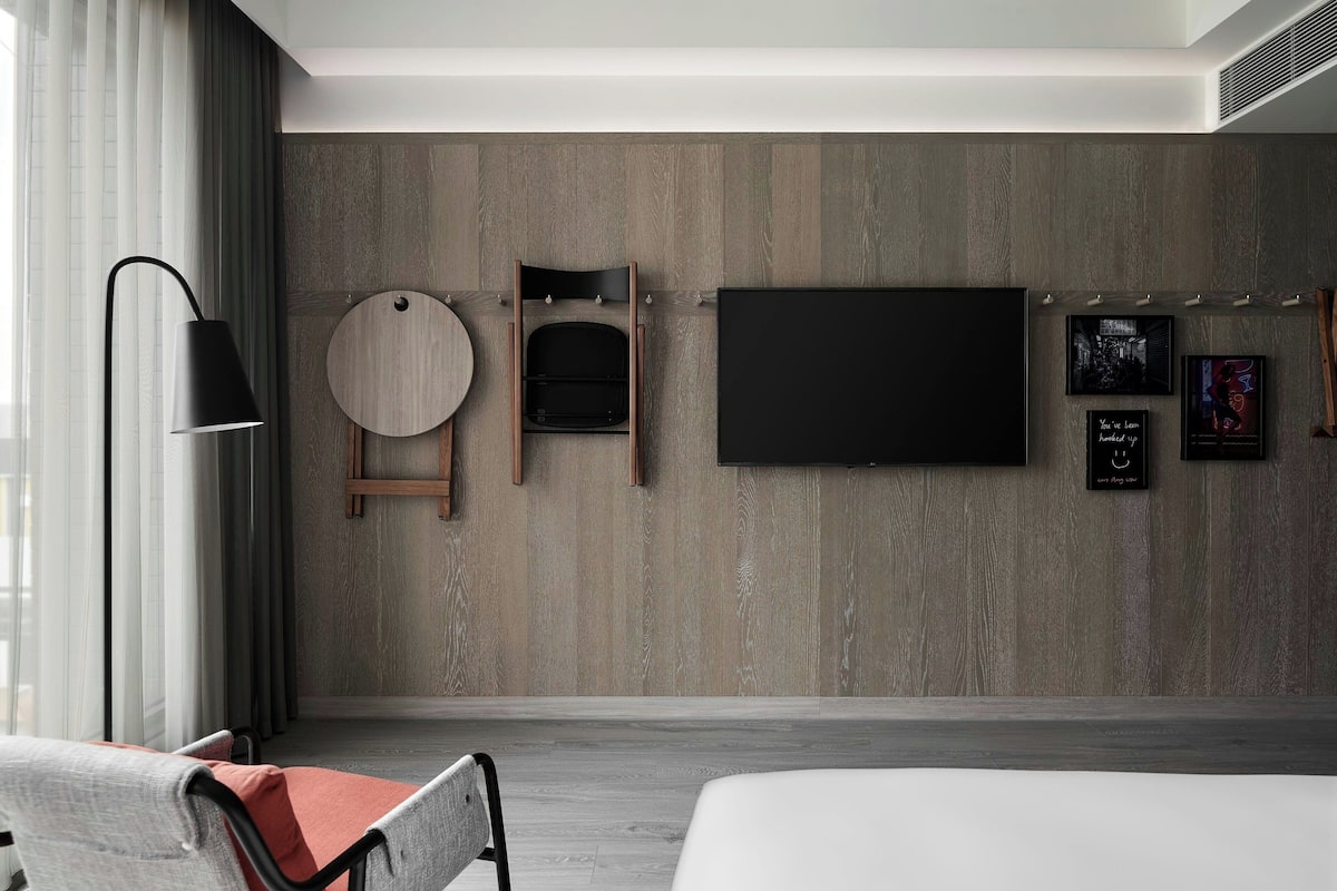 A modern hotel room with a large bed, a flat-screen TV mounted on the wall, a round wooden side table, a wooden chair, and a floor lamp.