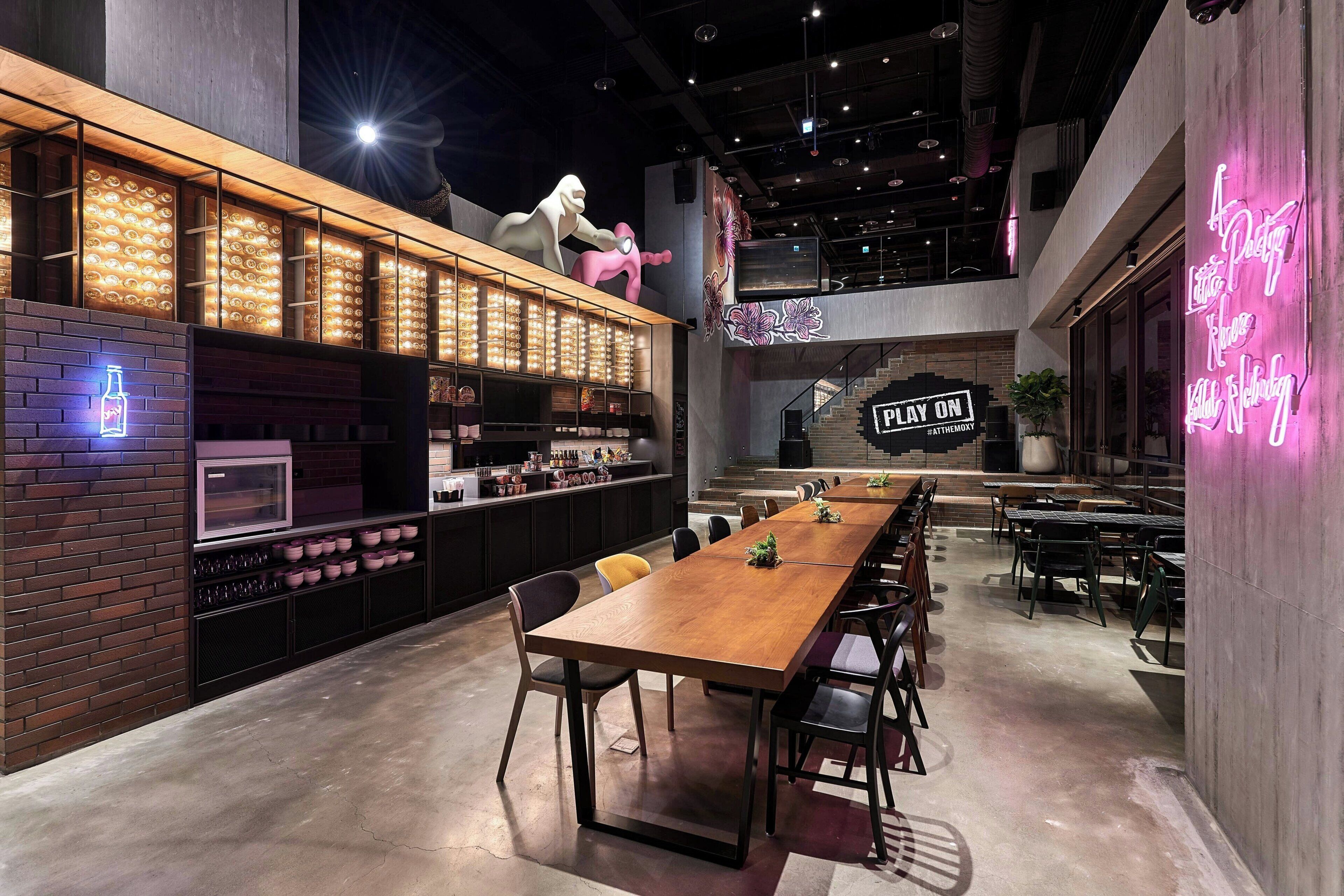 A modern restaurant with a long wooden table, black chairs, and a neon sign.