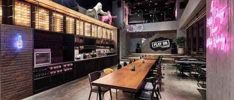 A modern restaurant with a long wooden table, black chairs, and a neon sign.