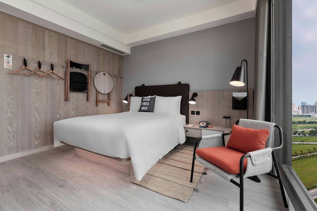 A modern hotel room with a large bed, a chair with a cushion, a nightstand with a lamp, and a view of the city through a large window.