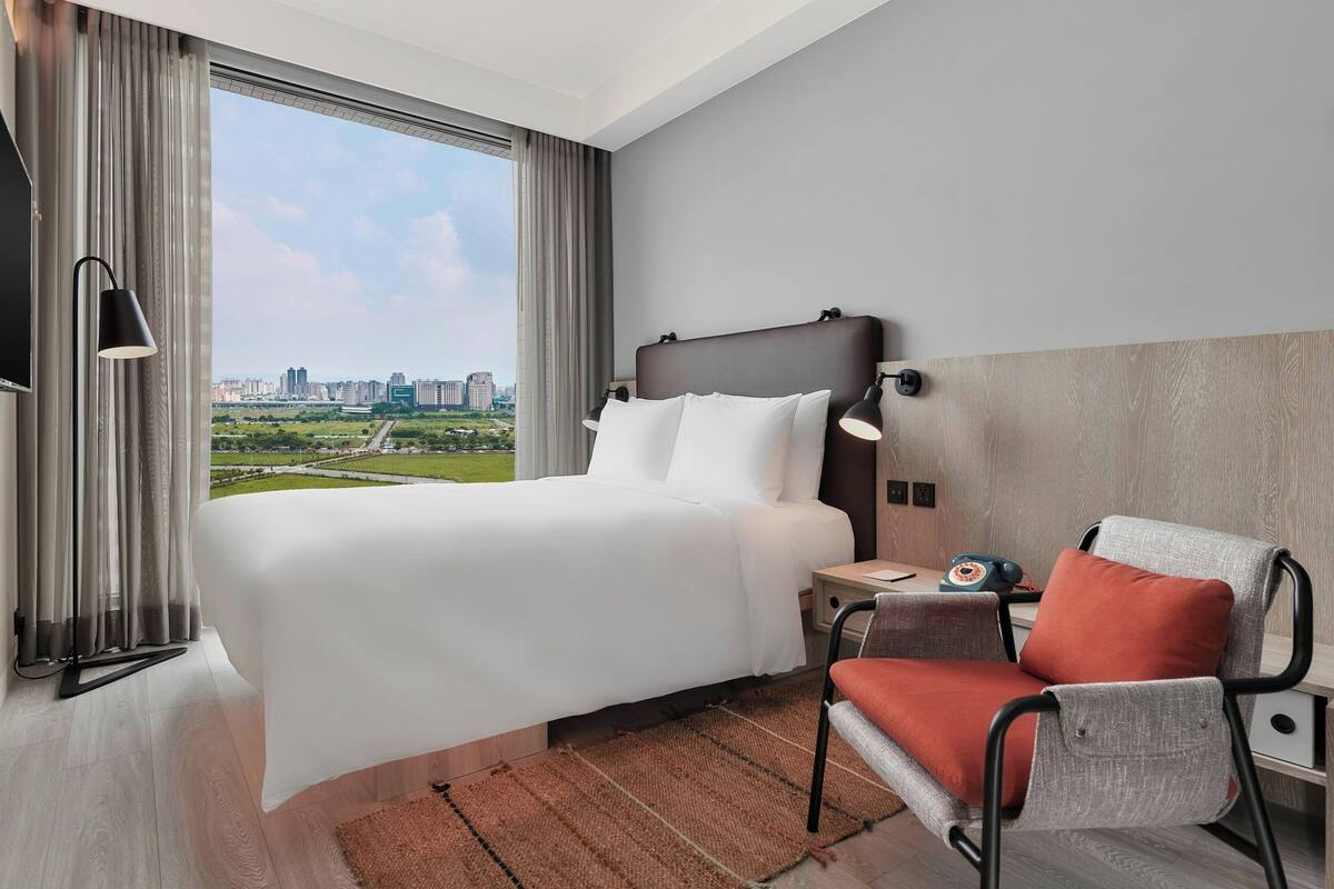 A hotel room with a large bed, a chair, a nightstand, and a view of a cityscape.