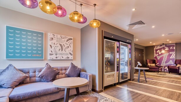 Un hall d’hôtel moderne avec un canapé, une table ronde, un pouf, des œuvres d’art décoratives au mur et un réfrigérateur.