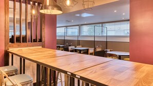 Un espace de restauration moderne avec des tables et des chaises en bois, des luminaires suspendus et de grandes fenêtres équipées de stores.